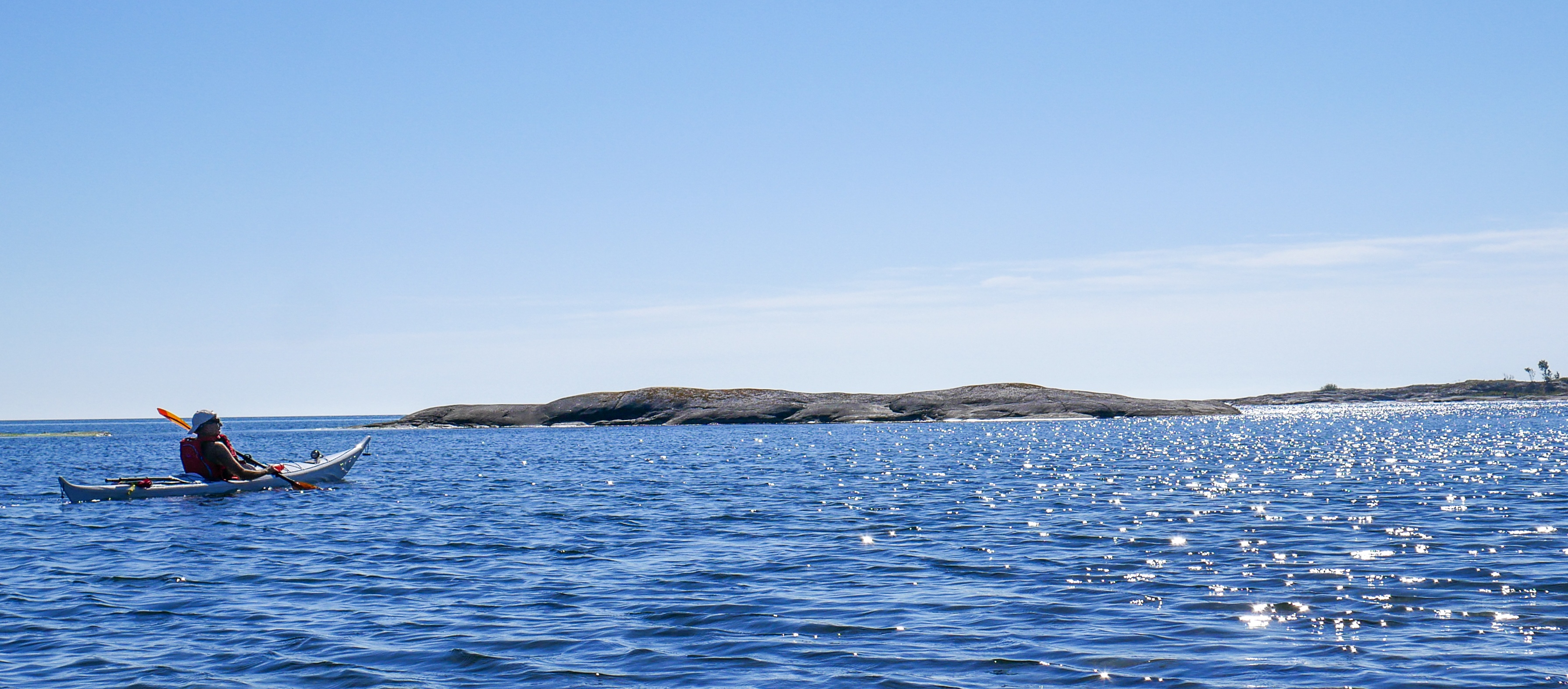 Kajakpaddling i öppet hav En person paddlar kajak i öppet hav under klarblå himmel med avlägsna öar i bakgrunden.