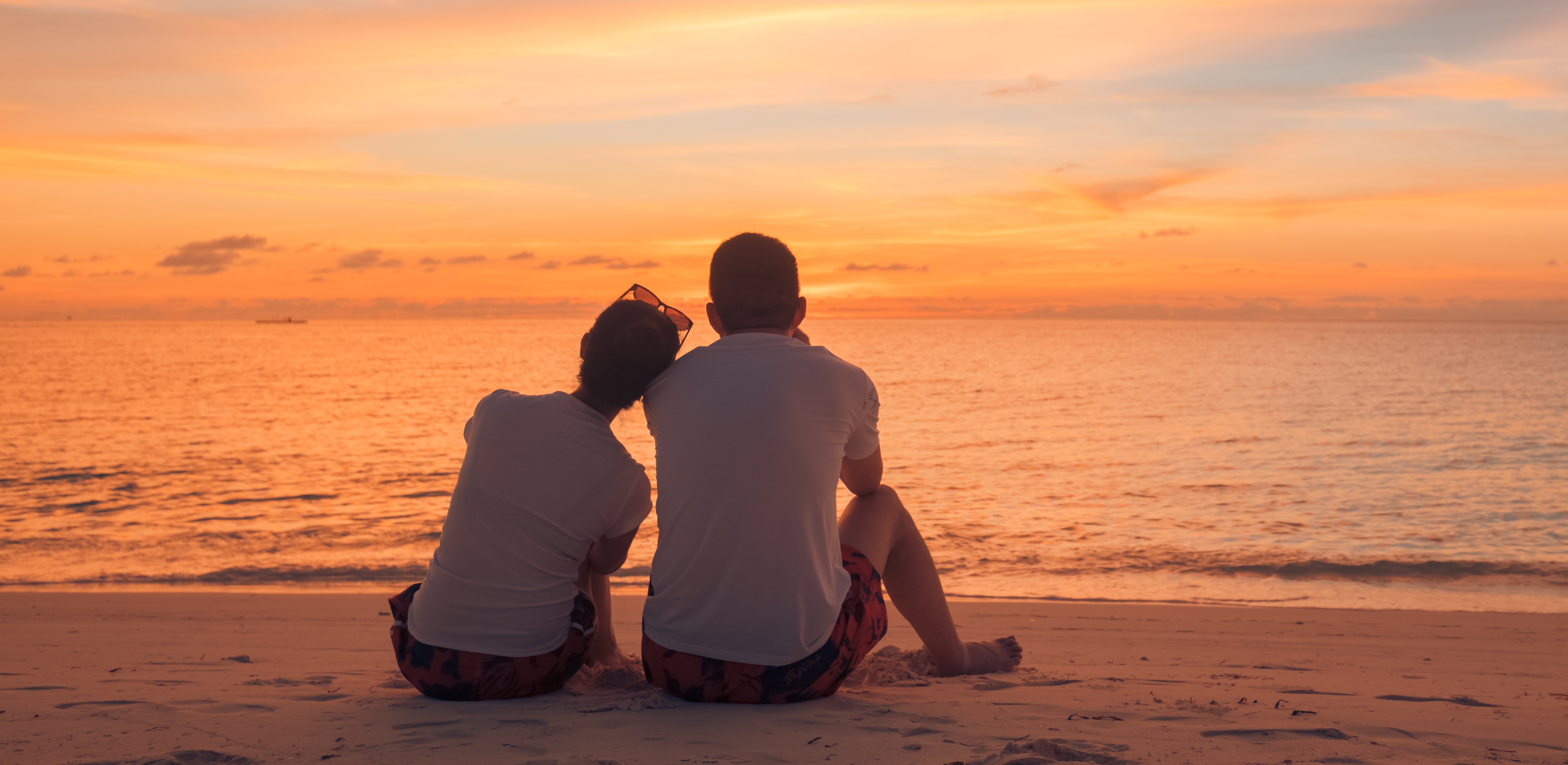 Par vid solnedgången Två personer sitter på stranden och tittar mot havet vid solnedgången. Himlen är färgad i orange och blå.