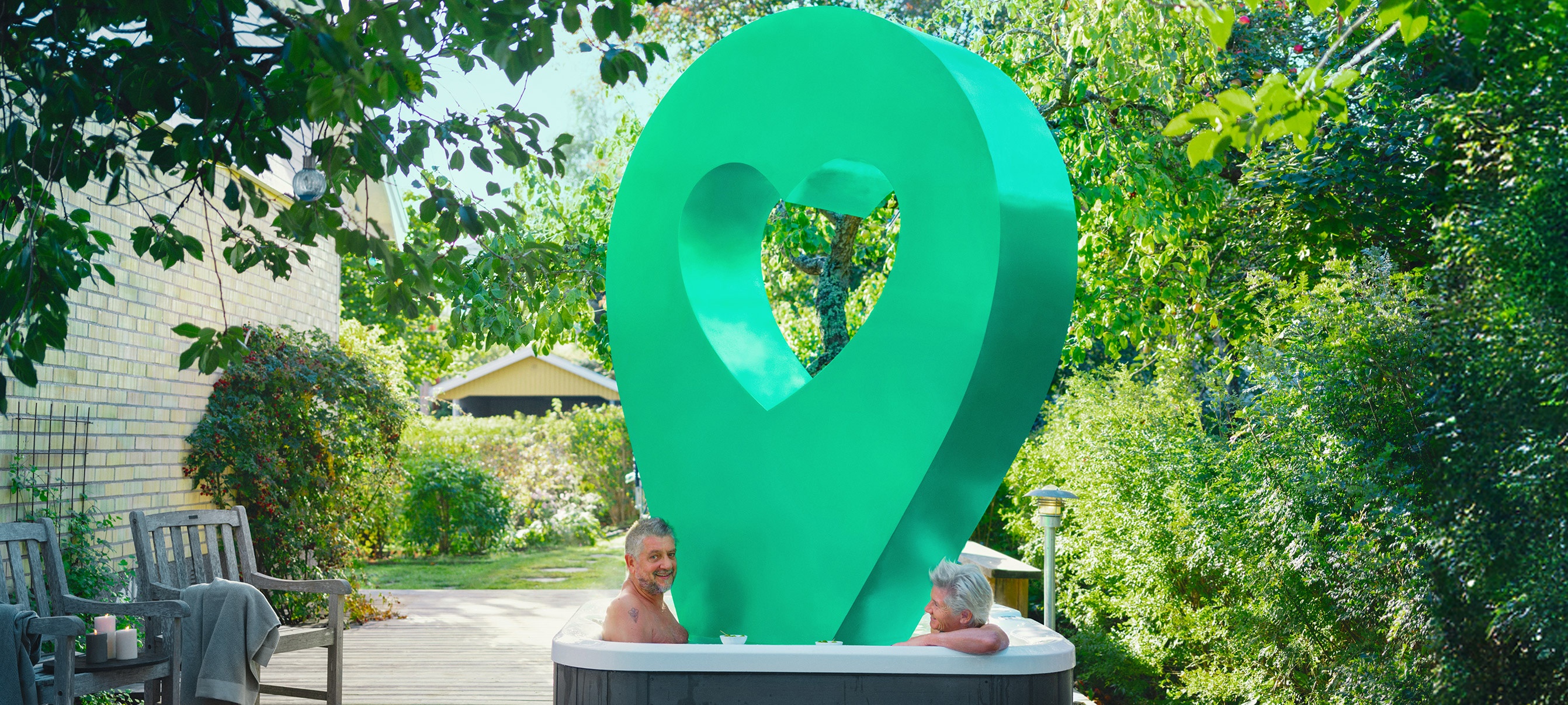 Bubbelpool utomhus Två personer sitter i en bubbelpool utomhus, omgiven av grönska och ett grönt hjärtformat konstverk.