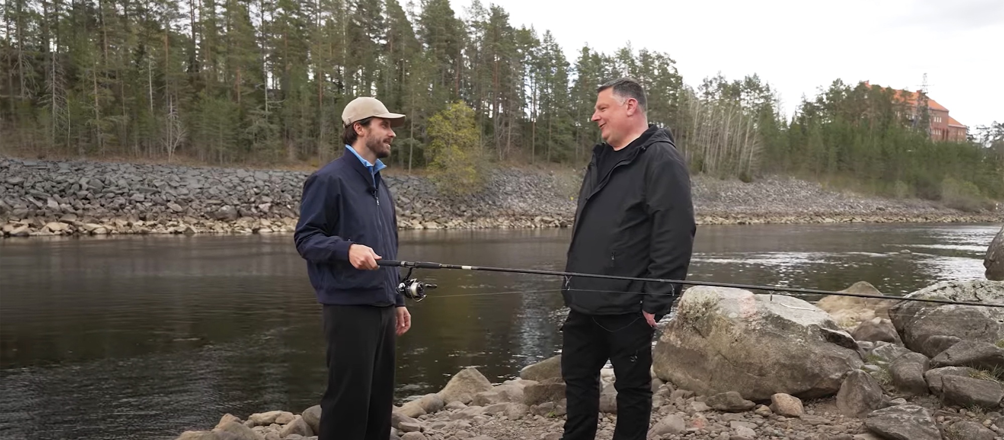 Victor och Christian vid Norören Två män står vid en flod och håller i ett fiskespö, omgiven av skog och stenar.