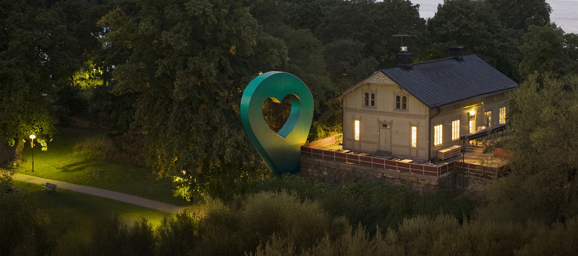 Lyckplatsen med hjärtskulptur Bild av Lyckplatsen som visar ett hus med ett hjärtaformad skulptur och vacker utsikt mot vattnet i skymningen.
