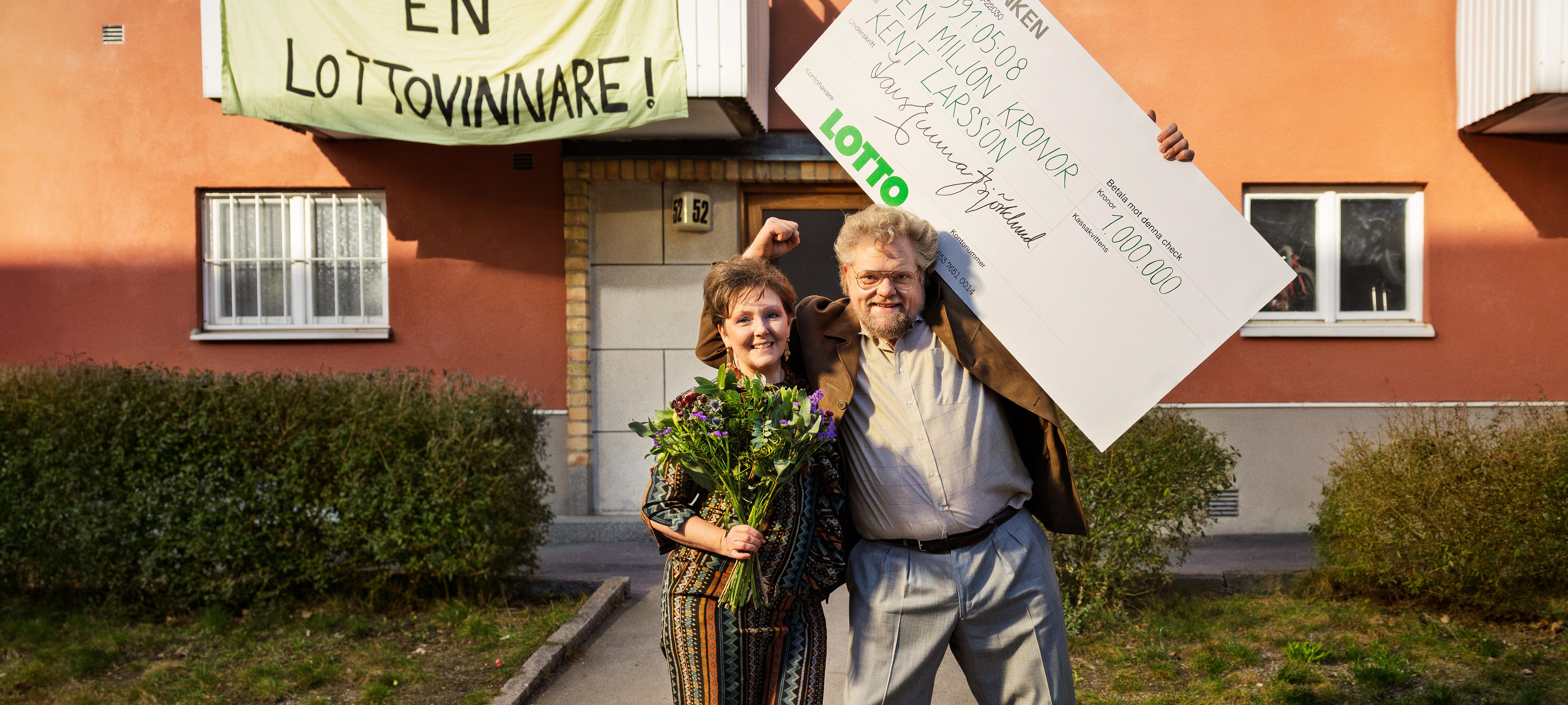 Glädje vid lottovinst Två personer firar med blommor och stor vinstcheck framför en byggnad med banderoll om lottovinst.