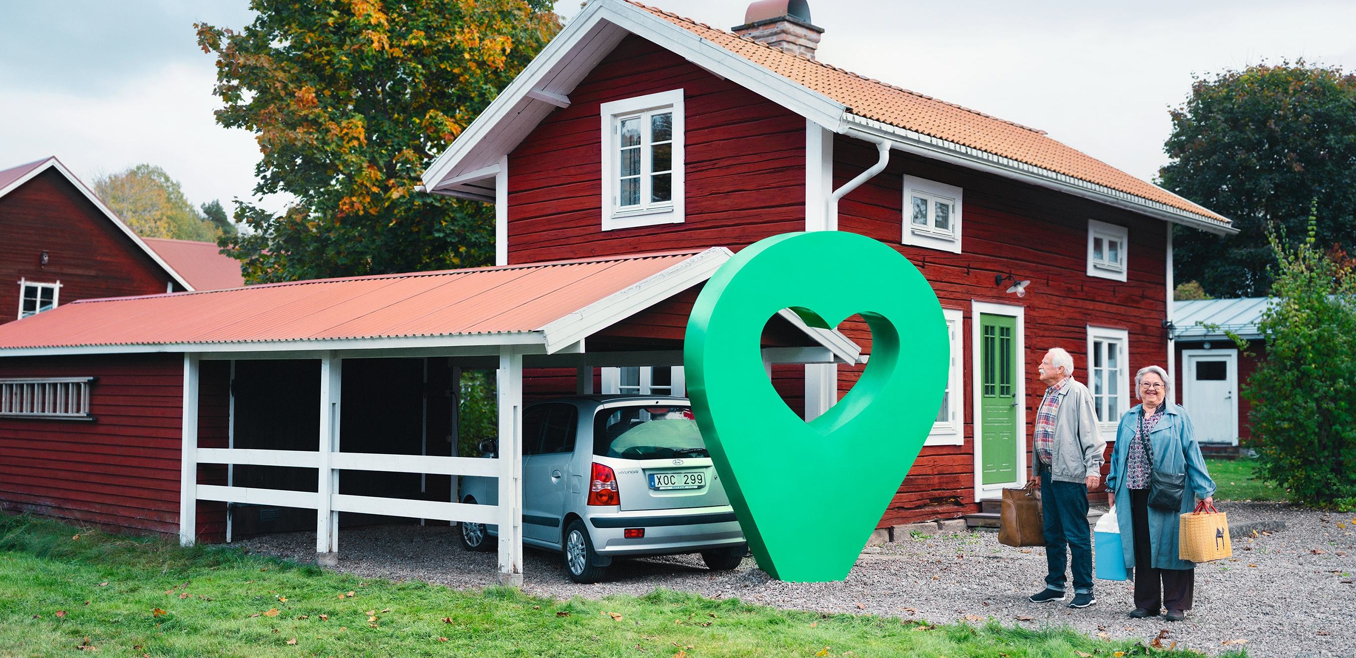 Rött hus med grönt hjärta Ett rött sommarhus med carport och två glada personer. En grön pin med hjärta är placerad vid huset.