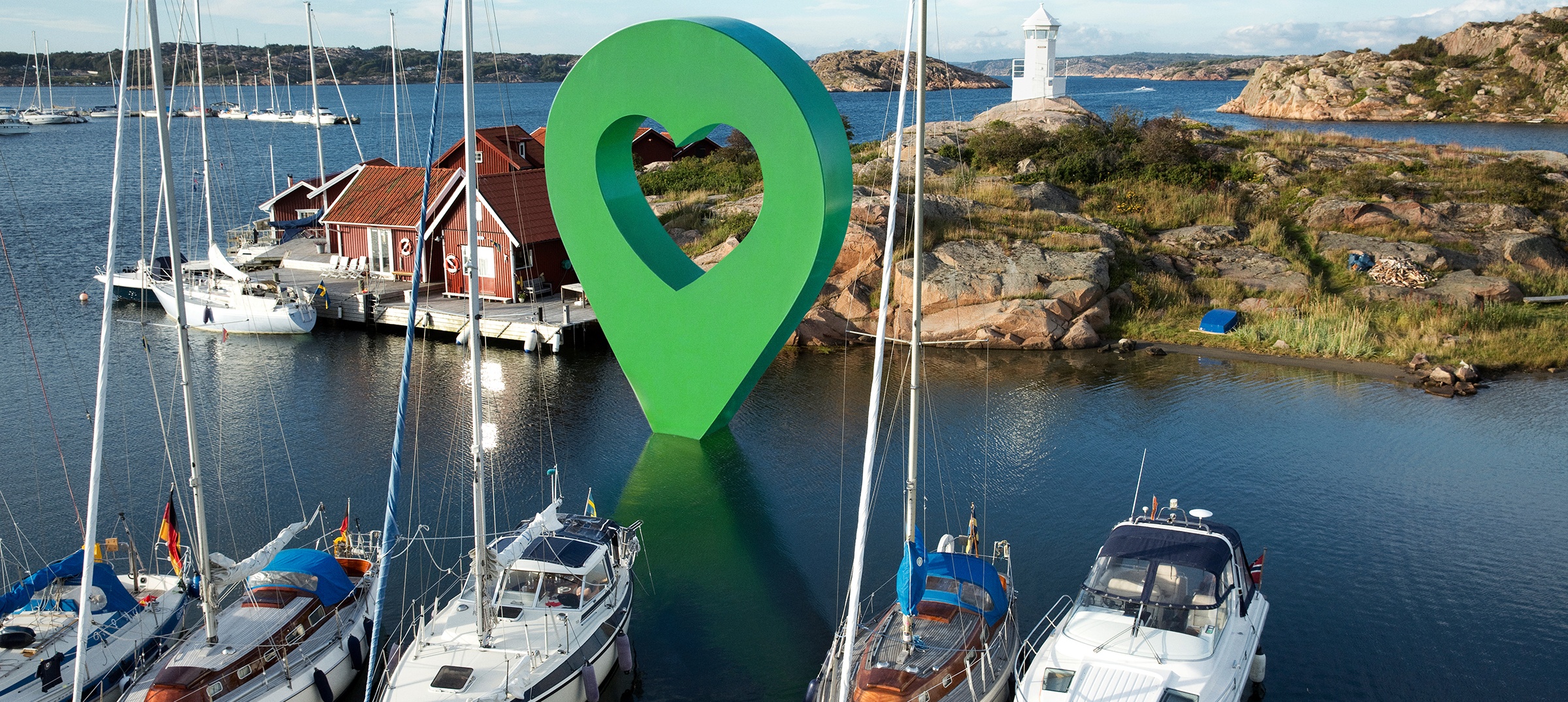 Svensk småbåtshamn med Lyckoplatsen-pin. Panoramavy av en svensk småbåtshamn med båtar och hus vid vattnet. En stor Lyckplatsen-pin sitter i viken.