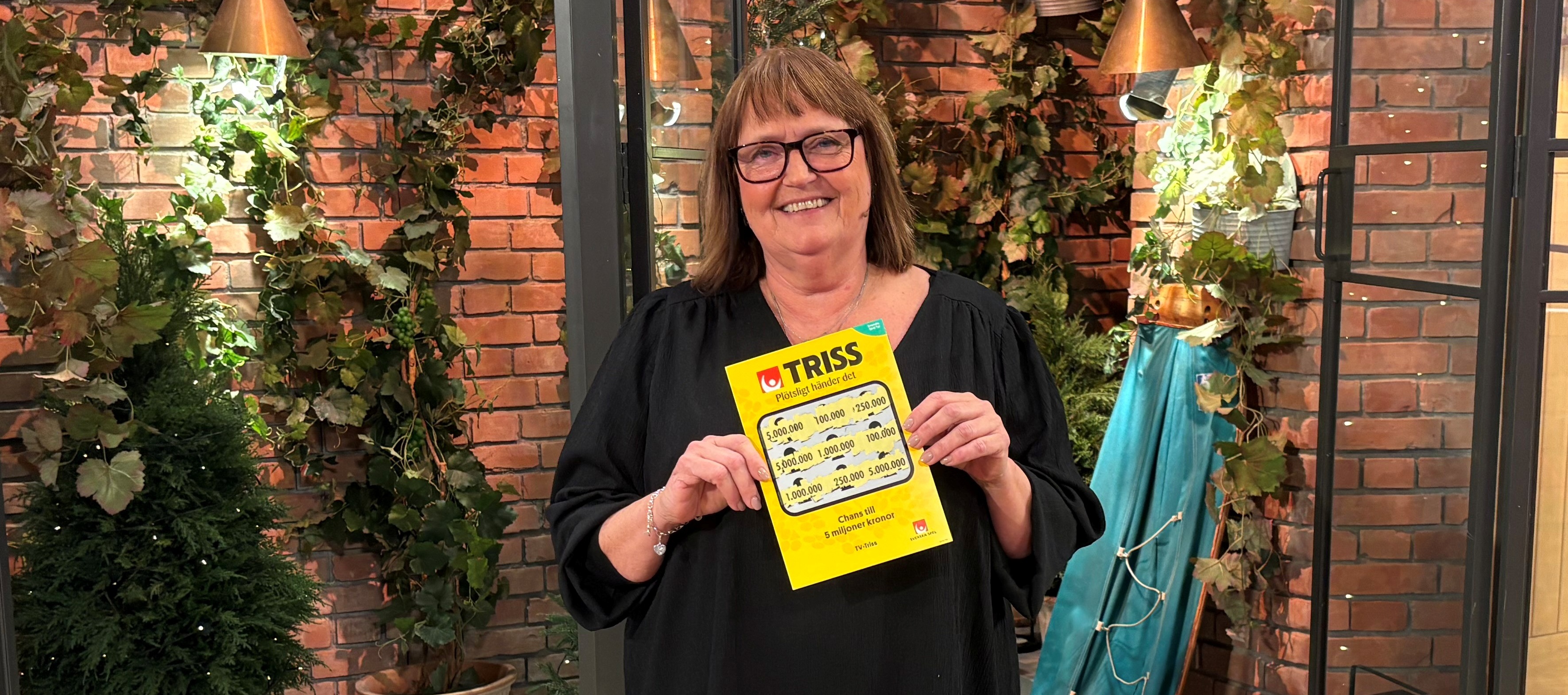 Person håller en Trisslott framför en dekorativ vägg med växter i bakgrunden.