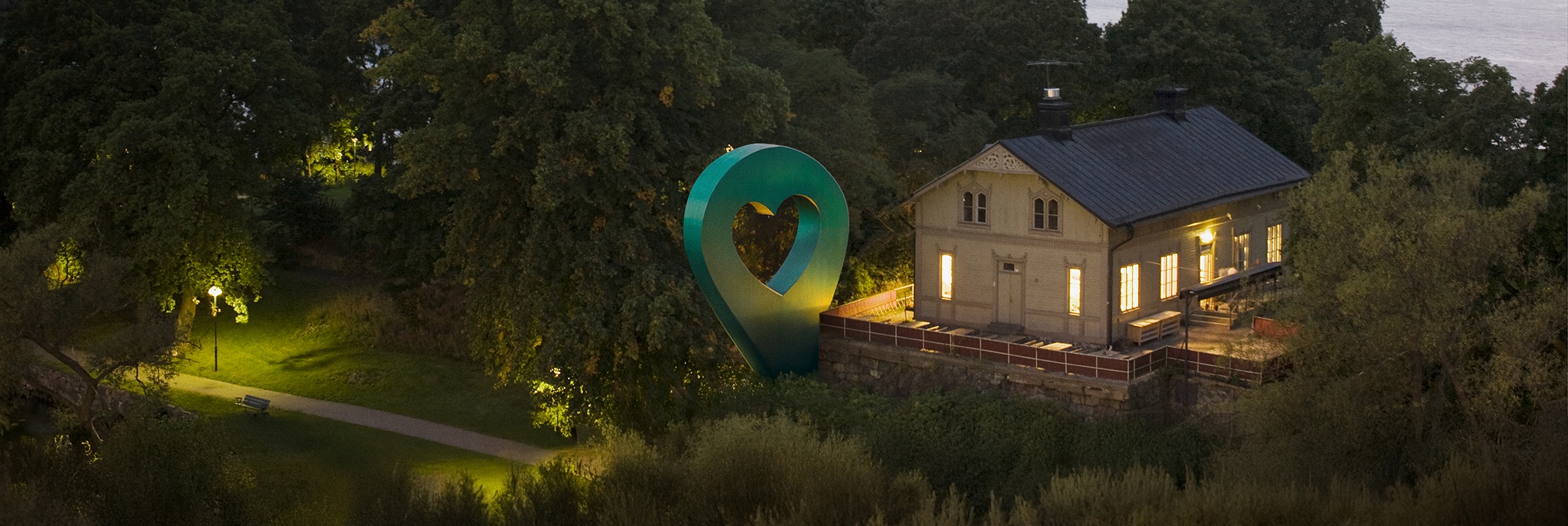 Bild av Lyckplatsen som visar ett hus med ett hjärtaformad skulptur och vacker utsikt mot vattnet i skymningen.