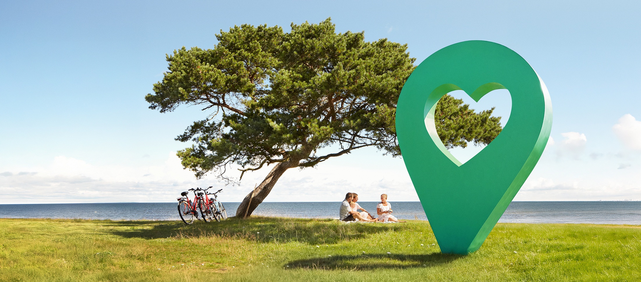 En vacker sommardag med blå himmel, människor sitter på gräset under ett träd bredvid en grön lyckoplatsmarkör.