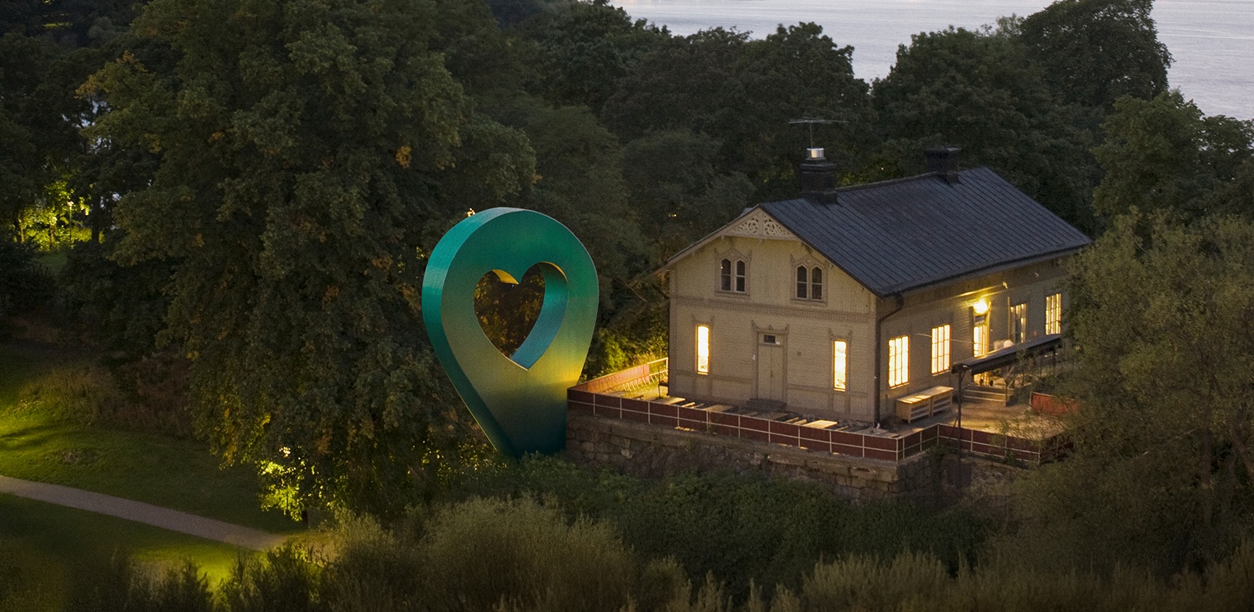 Bild av Lyckplatsen som visar ett hus med ett hjärtaformad skulptur och vacker utsikt mot vattnet i skymningen.