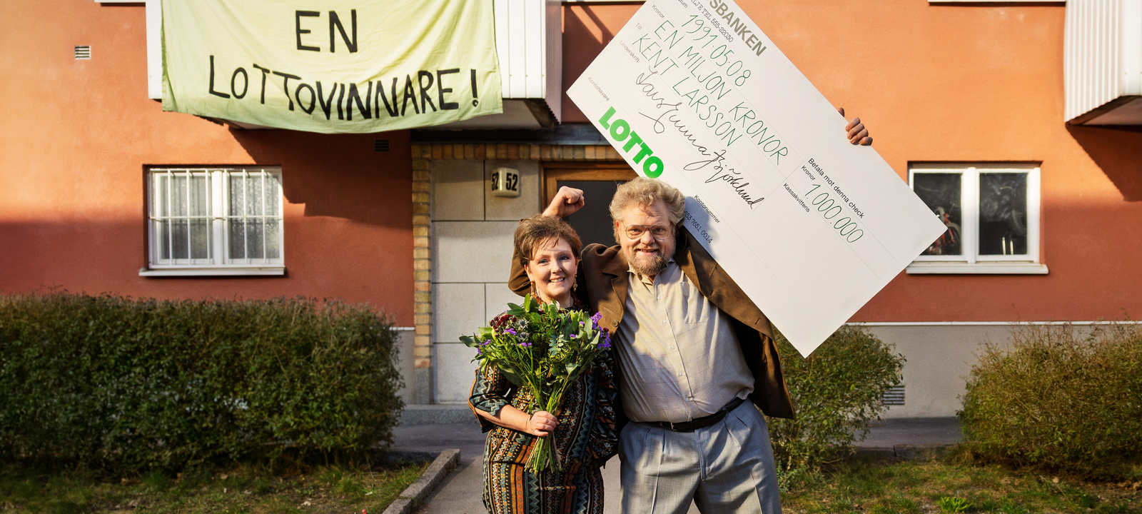 Två personer firar med blommor och stor vinstcheck framför en byggnad med banderoll om lottovinst.