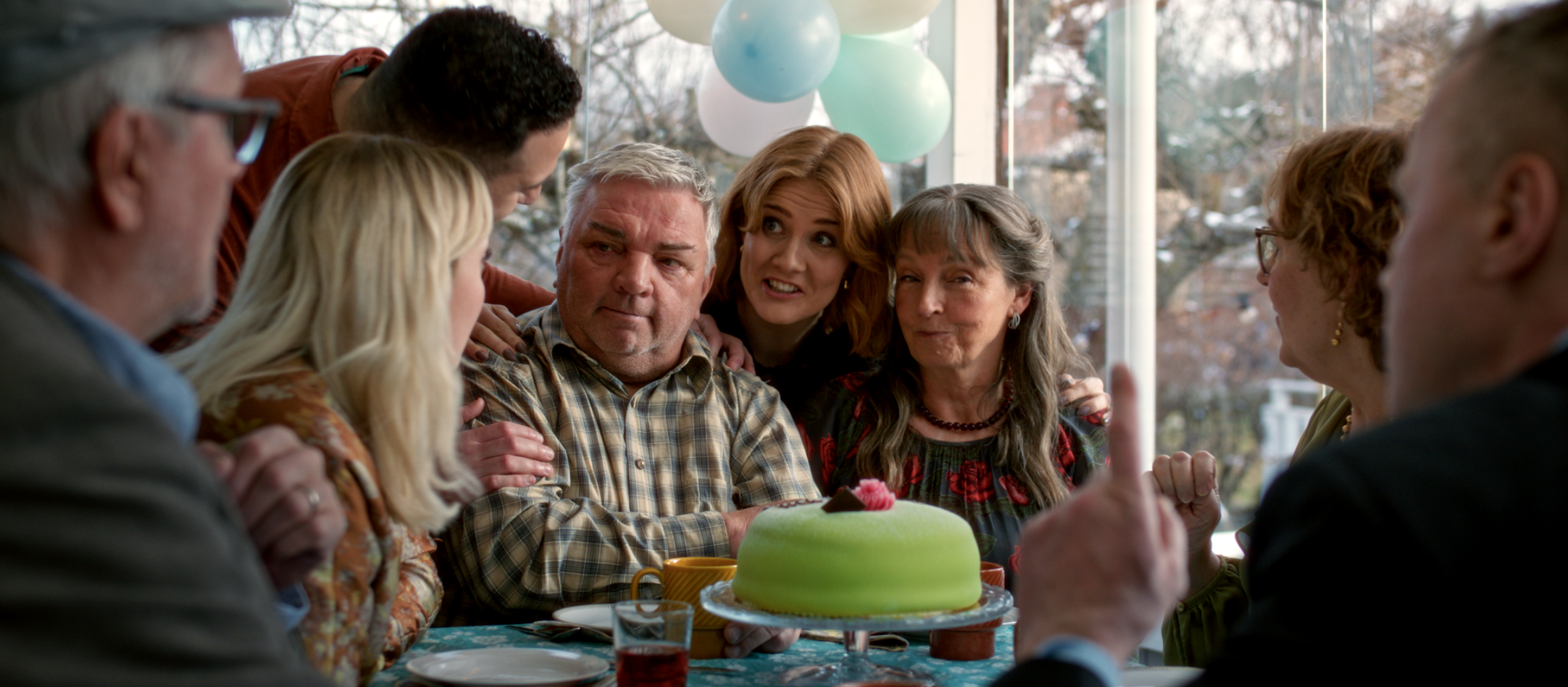 En grupp människor samlade runt ett bord med en grön tårta, ballonger i bakgrunden.