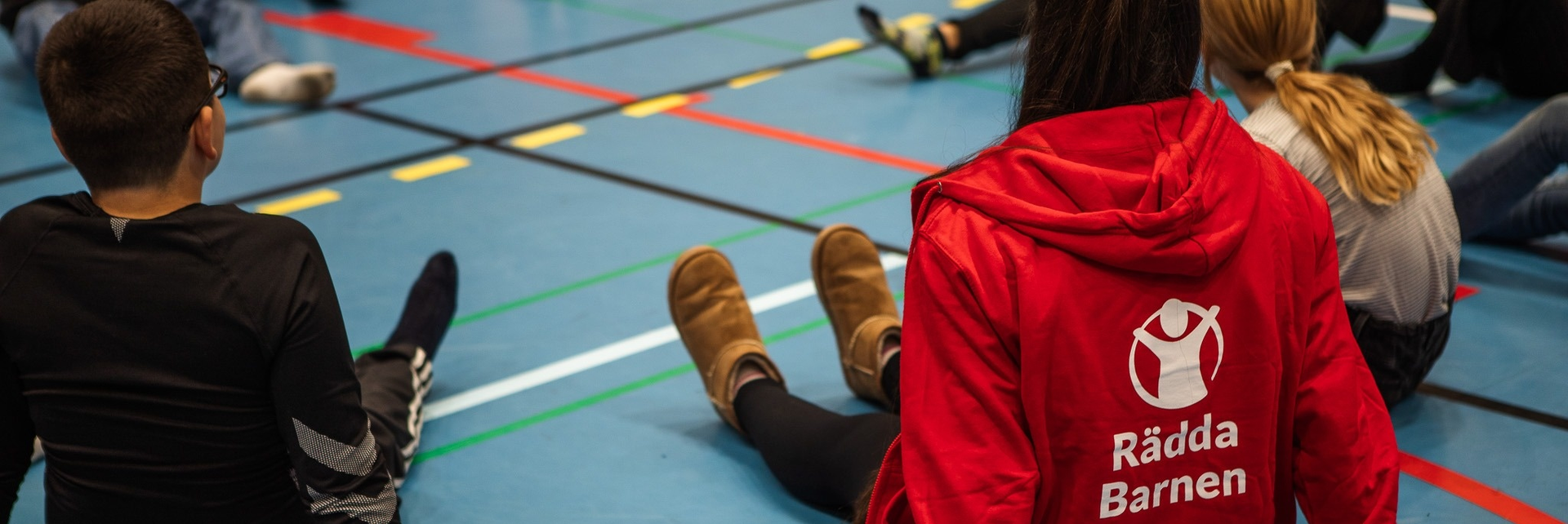 Personer sitter på ett golv i en gympasal, en har på sig en röd Rädda Barnen-jacka.
