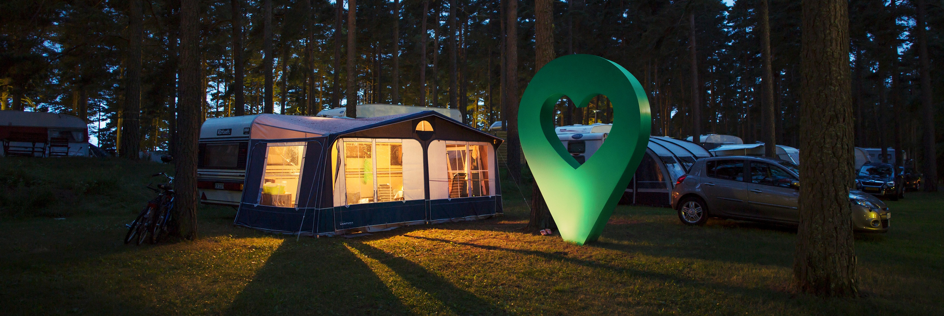 Kvällsbild på en tältplats i en skog med en stor grön markör som lyser. Campingtältet är upplyst inifrån.