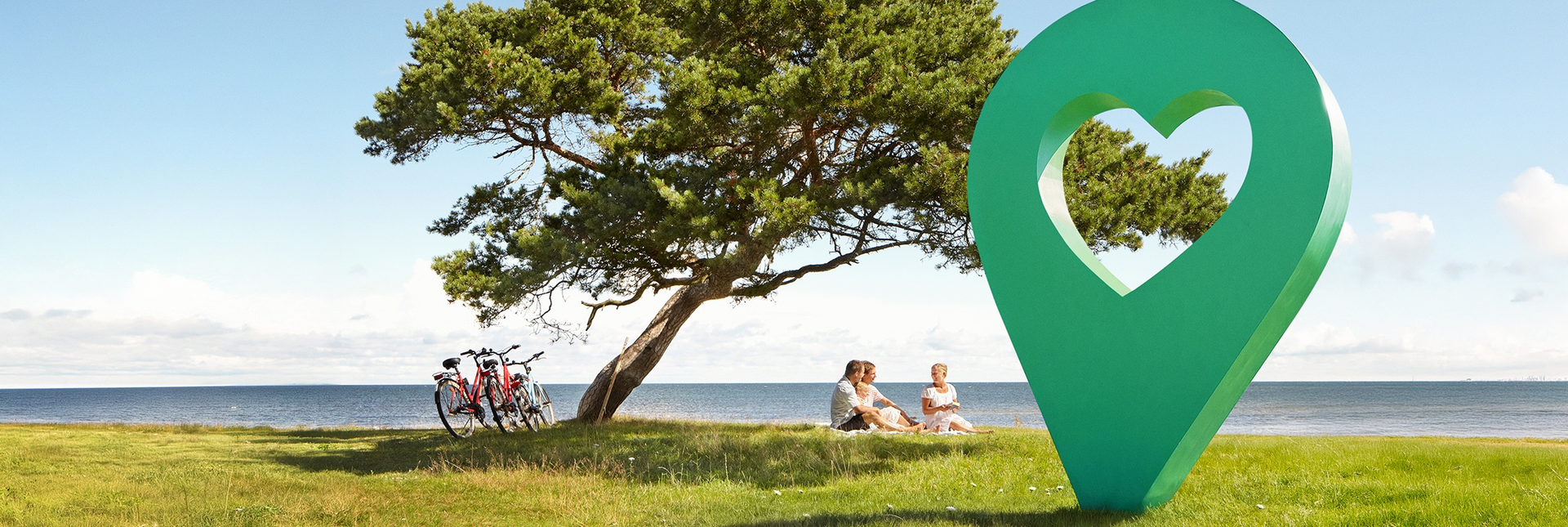 En vacker sommardag med blå himmel, människor sitter på gräset under ett träd bredvid en grön lyckoplatsmarkör.