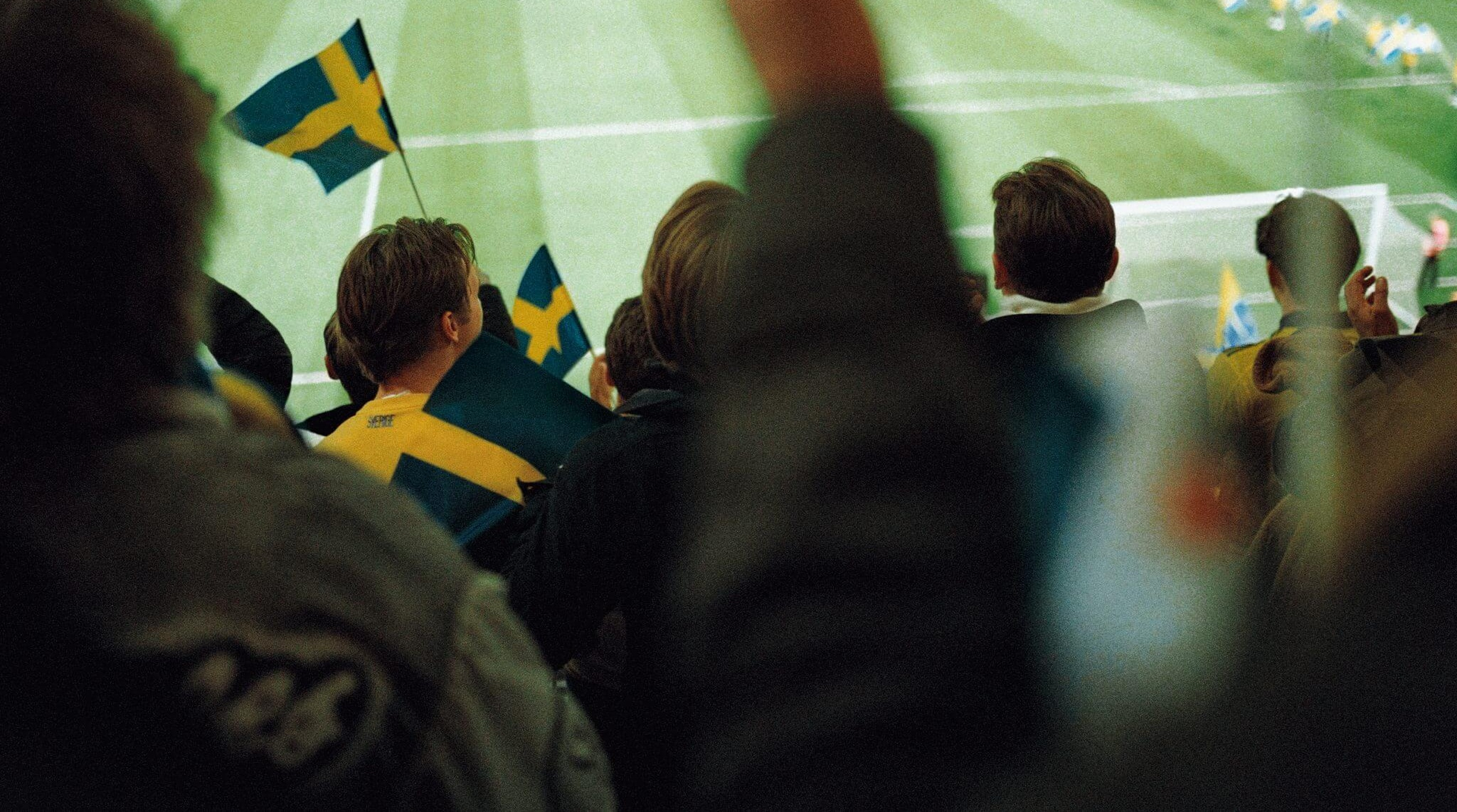 Publik på läktaren viftar med svenska flaggor och en fotbollsplan avtecknar sig i bakgrunden.