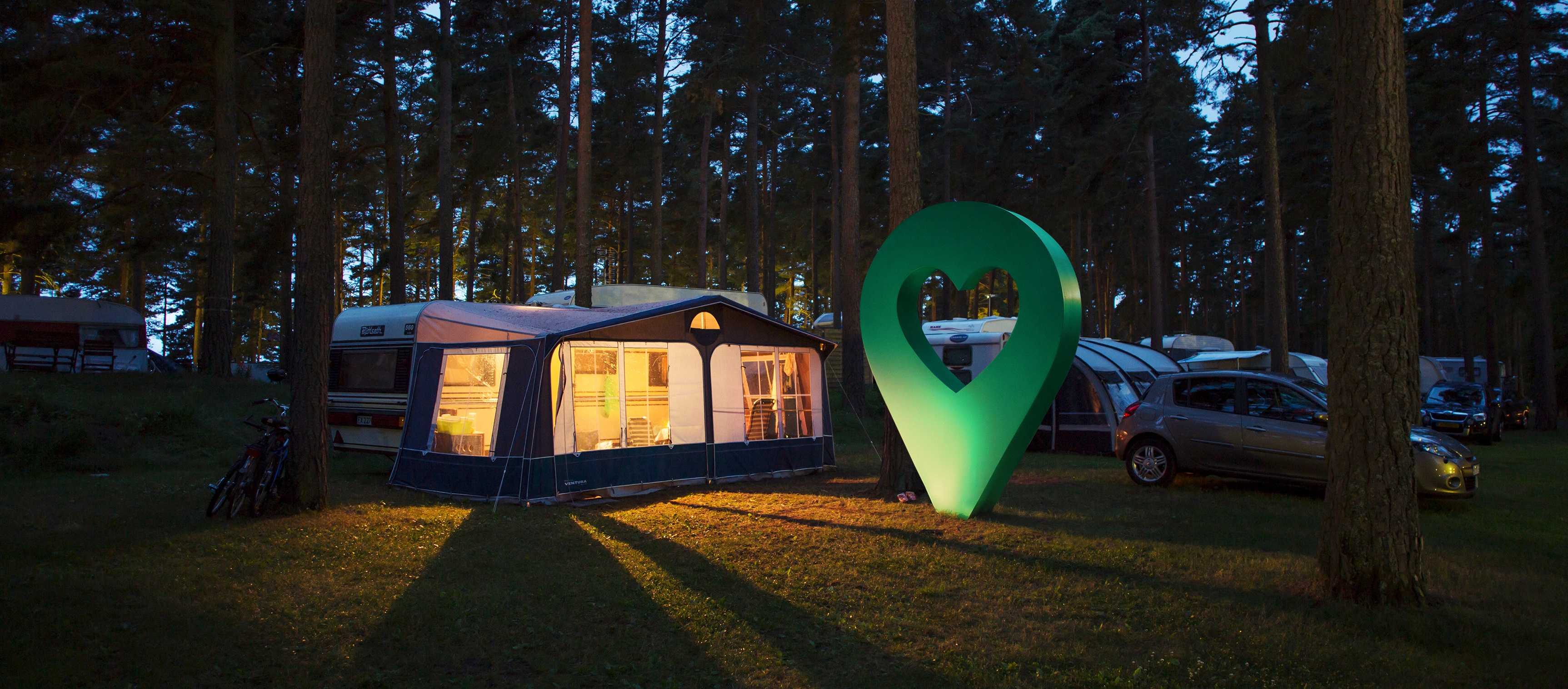 Kvällsbild på en tältplats i en skog med en stor grön markör som lyser. Campingtältet är upplyst inifrån.