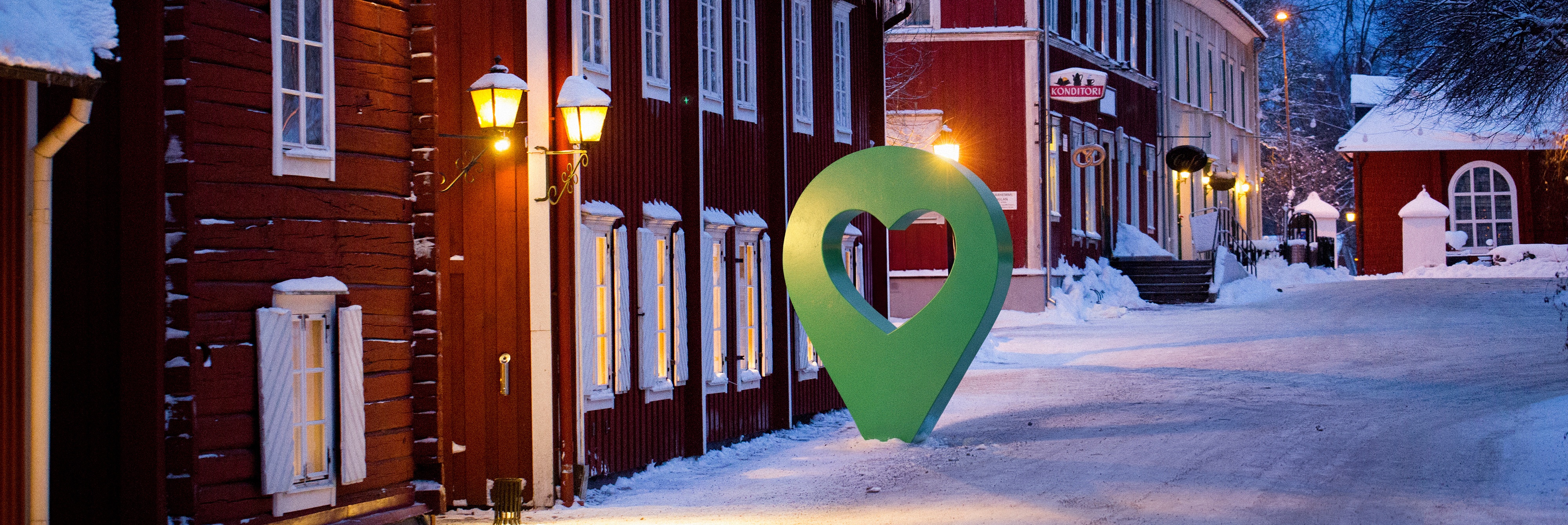 Vinterbild av en småstadsgata med röda trähus och en grön hjärtsymbol intill en lyktstolpe.
