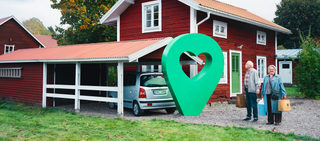 Ett rött sommarhus med carport och två glada personer. En grön pin med hjärta är placerad vid huset.