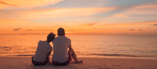 Två personer sitter på stranden och tittar mot havet vid solnedgången. Himlen är färgad i orange och blå.