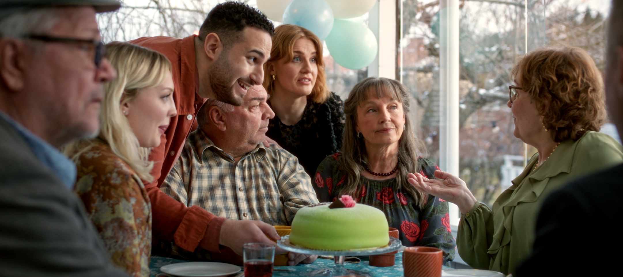 En grupp människor sitter runt ett bord och tittar på en person som håller i en grön tårta.