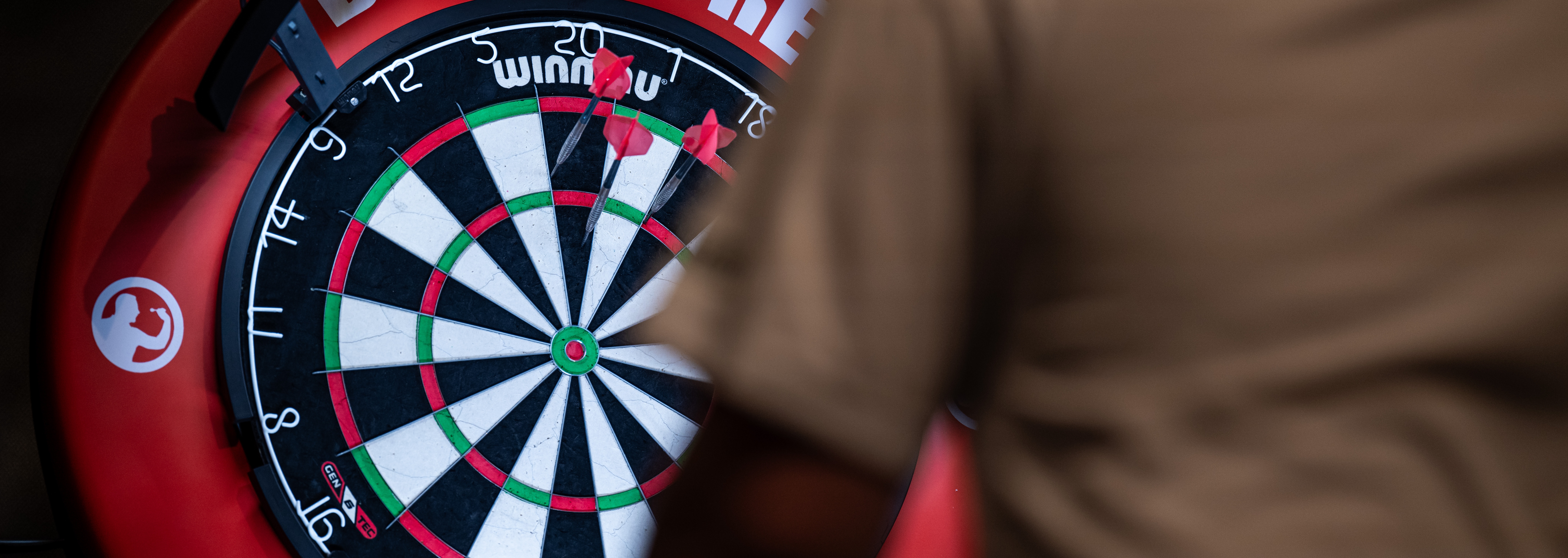 En person kastar pil mot en darttavla med röd ram och vitt varumärke synligt.