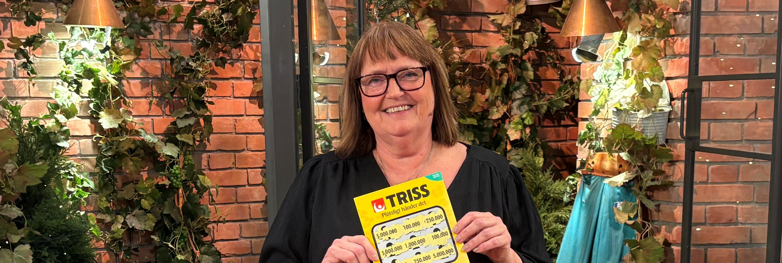 Person håller en Trisslott framför en dekorativ vägg med växter i bakgrunden.