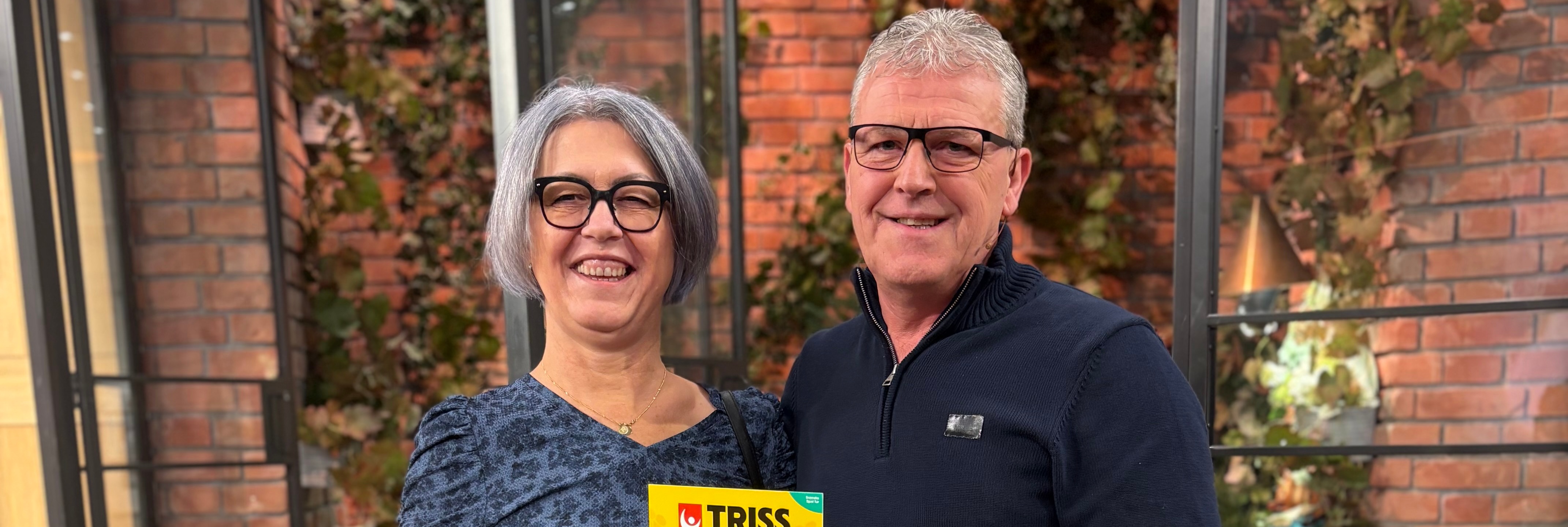 Två glada personer poserar med en gul Triss-lott framför en vägg av tegel och grönt lövverk.