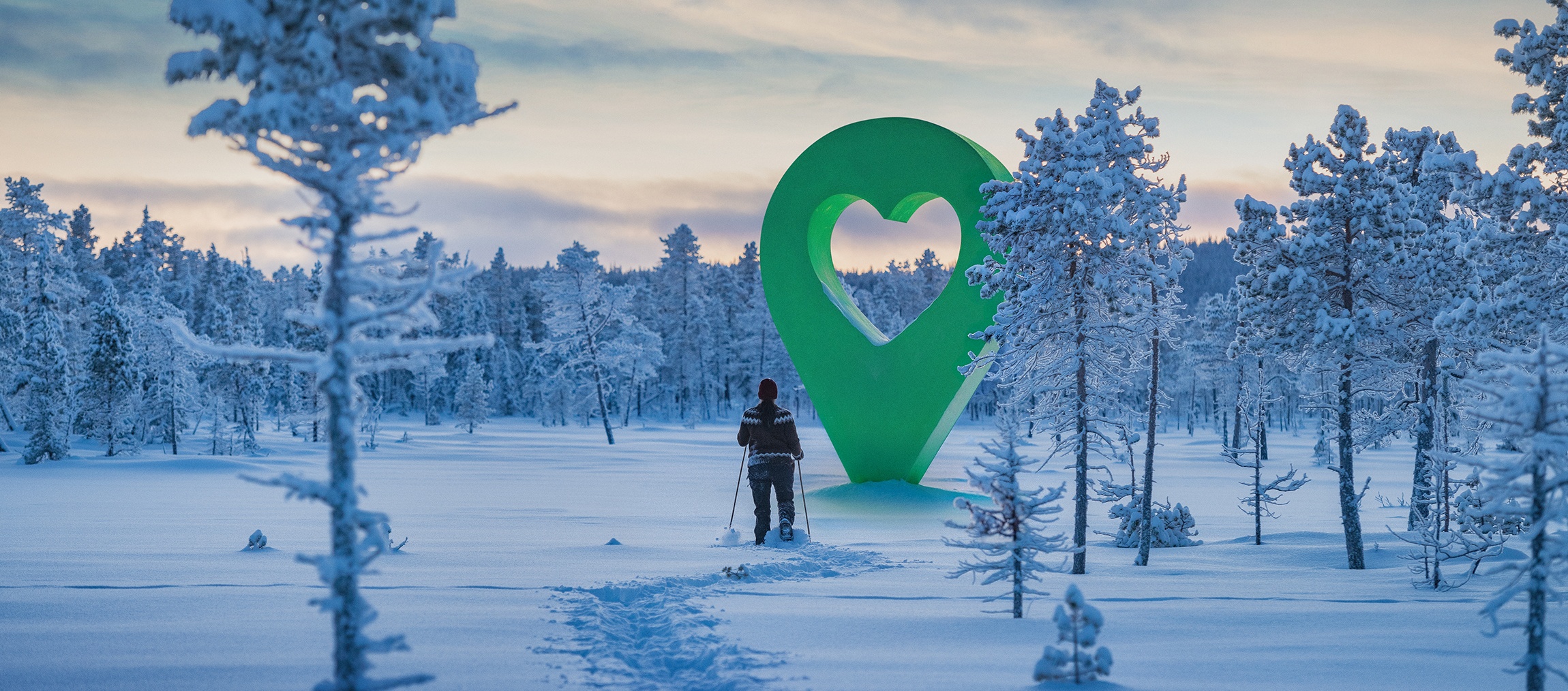 Person vandrar i vinterlandskap mot stor grön hjärtsymbol. Avbildar en vacker plats i naturen med snöklädda träd.