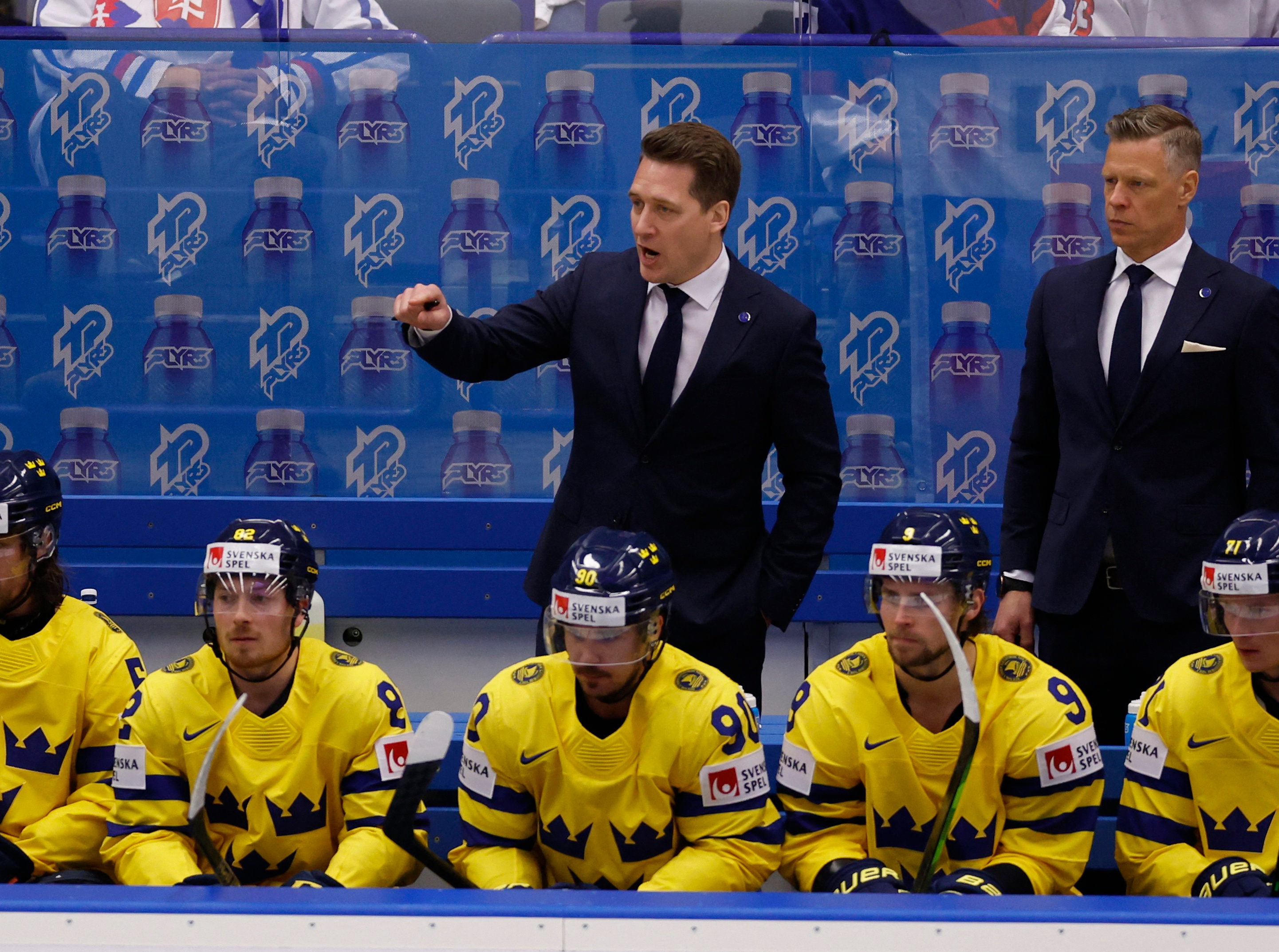 Svenska ishockeylaget Tre Kronor i sina gula tröjor på bänken under match, två tränare står bakom.
