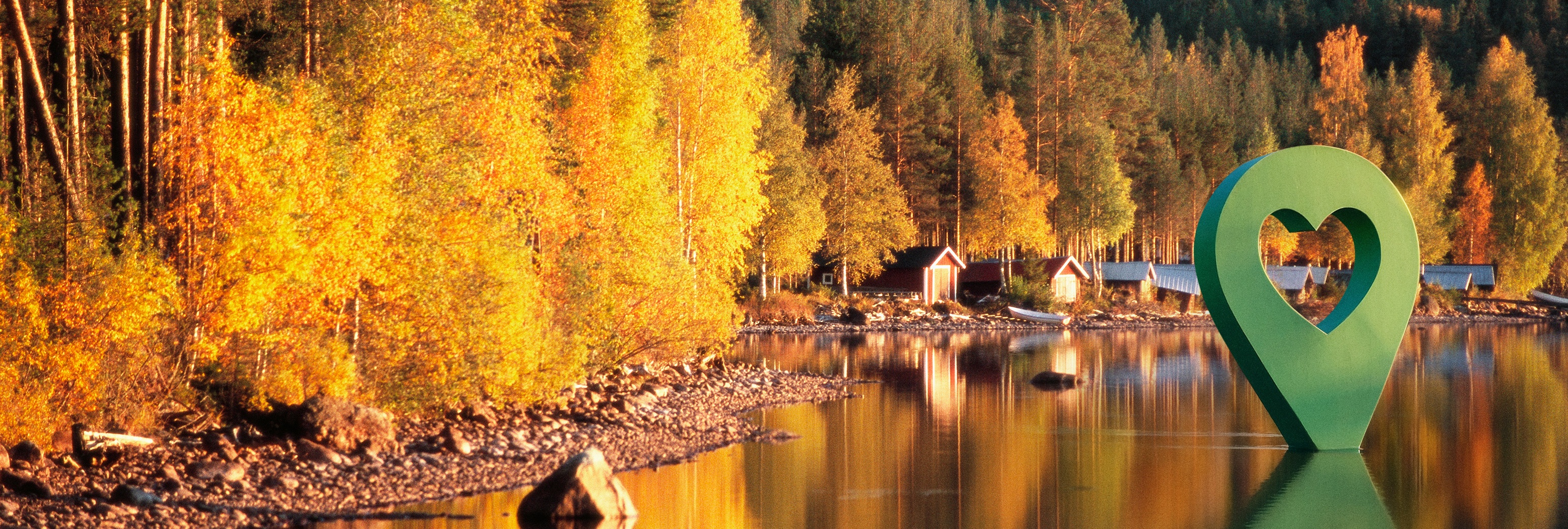 Bild av Lyckplatsen med höstfärger, omringad av träd och en grön hjärtformad markör vid sjön.