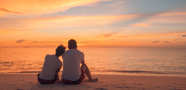 Två personer sitter på stranden och tittar mot havet vid solnedgången. Himlen är färgad i orange och blå.