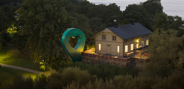 Bild av Lyckplatsen som visar ett hus med ett hjärtaformad skulptur och vacker utsikt mot vattnet i skymningen.