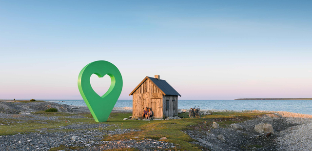 En liten stuga vid havet med en grön lyckoplatsmarkering i form av en hjärta placerad framför stugan.