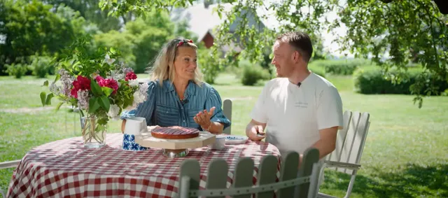 Gabriel Jonsson och Karin Almström sitter vid ett bord med rutig duk i en trädgård en solig dag. På bordet står en rabarberkaka.