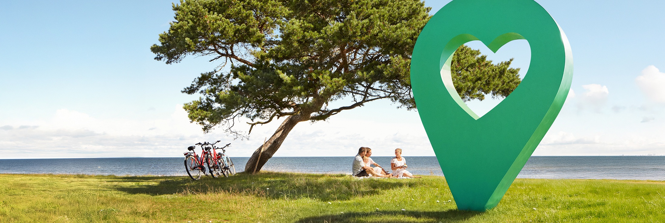 En vacker sommardag med blå himmel, människor sitter på gräset under ett träd bredvid en grön lyckoplatsmarkör.