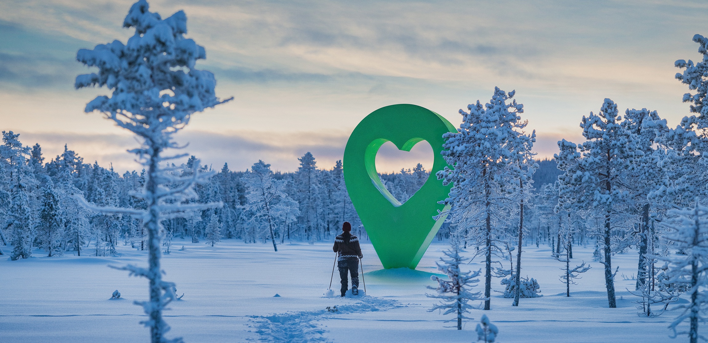 Person vandrar i vinterlandskap mot stor grön hjärtsymbol. Avbildar en vacker plats i naturen med snöklädda träd.