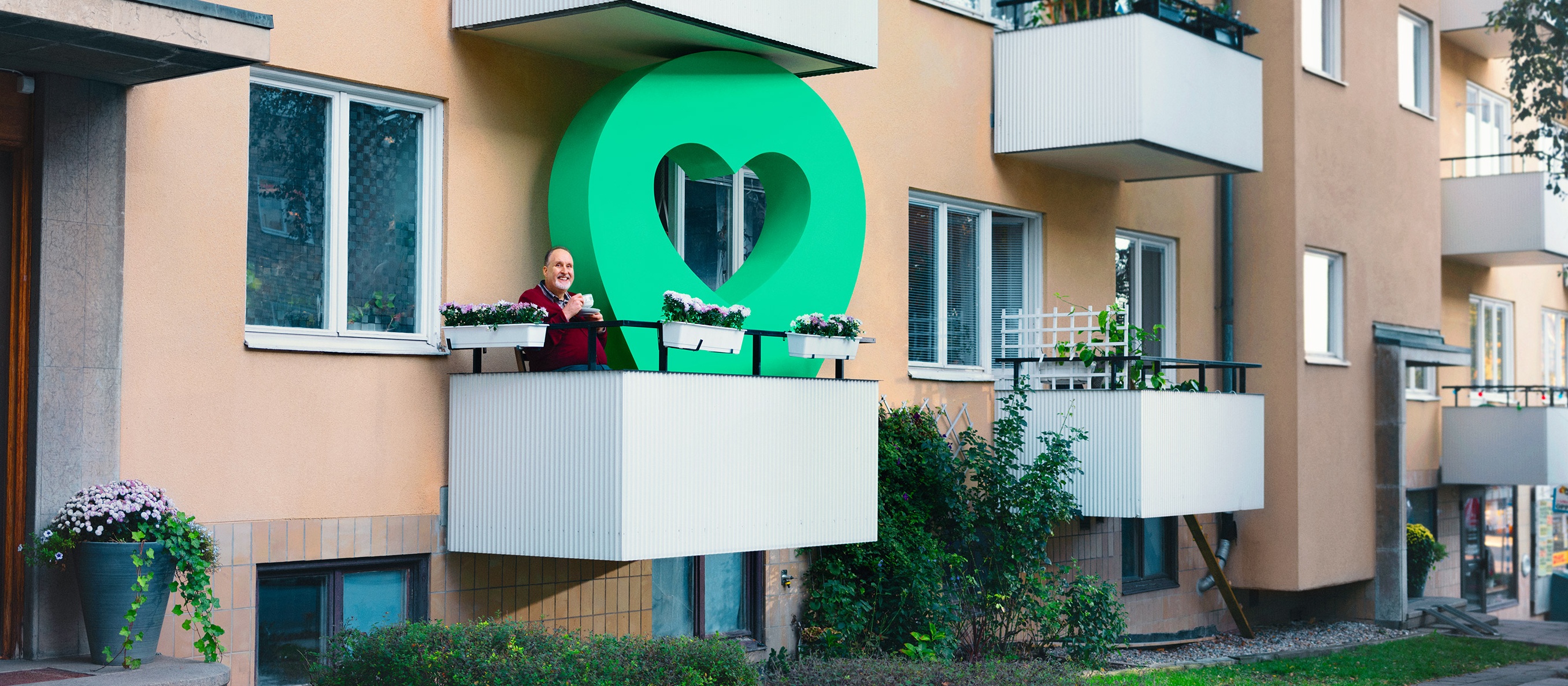 Man dricker kaffe på en balkong med en stor grön pin i ett blomsterfyllt lägenhetshusområde.