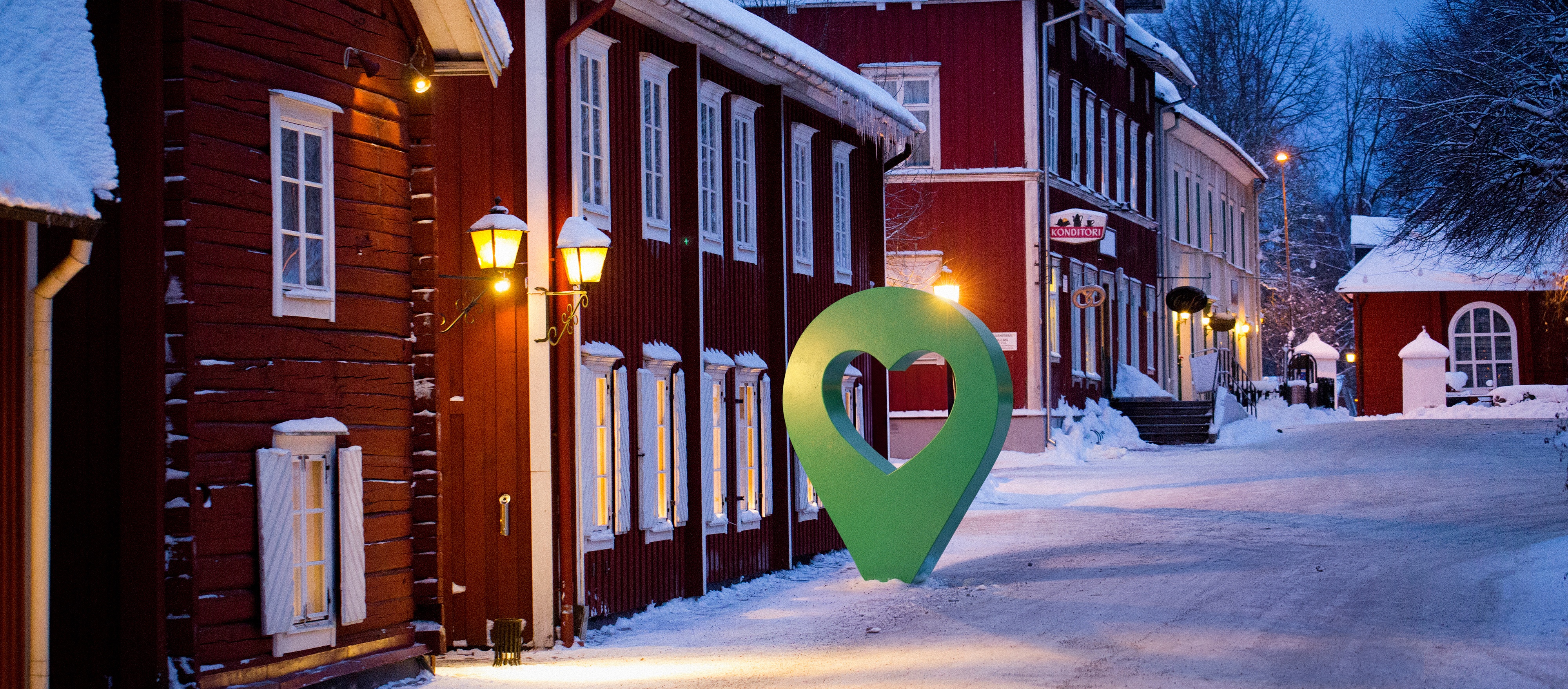 Vinterbild av en småstadsgata med röda trähus och en grön hjärtsymbol intill en lyktstolpe.