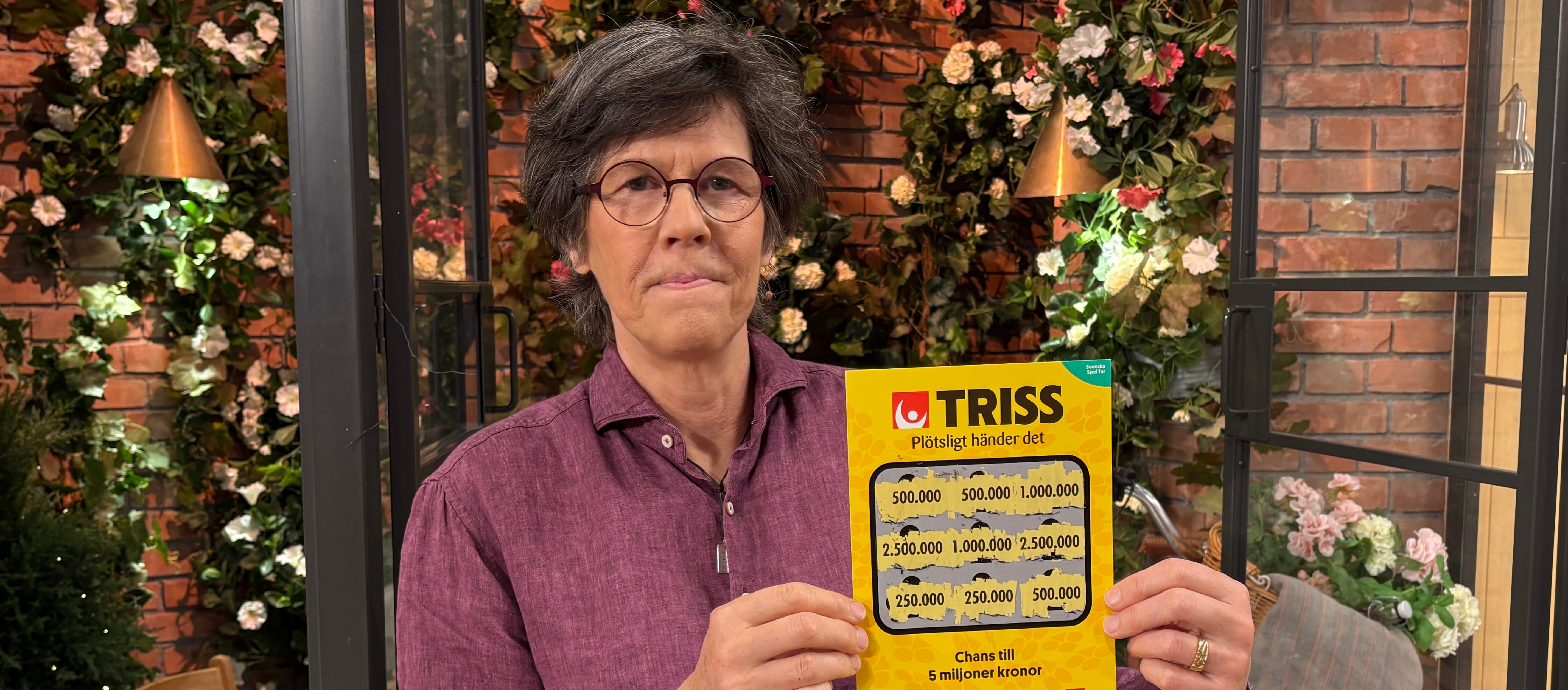 Person håller i en gul Trisslott framför en blomvägg. Chancen att vinna varierande summor pengar.