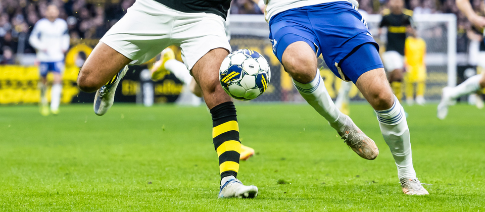 Två spelare i Allsvenskan sparkar mot bollen på en fotbollsarena, den ena i svart och den andra i vitt.