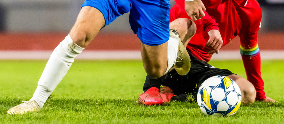 Två fotbollsspelare tävlar om bollen på gräsmatta, en i blå och en i röd tröja, under Superettan match.
