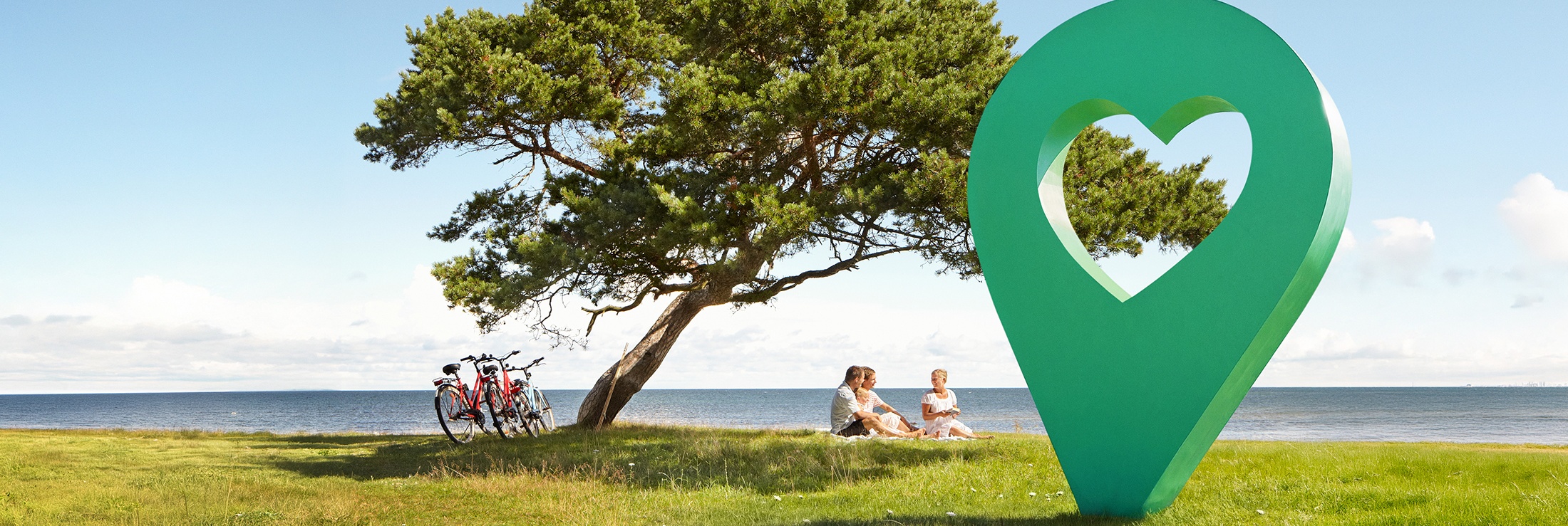 En vacker sommardag med blå himmel, människor sitter på gräset under ett träd bredvid en grön lyckoplatsmarkör.