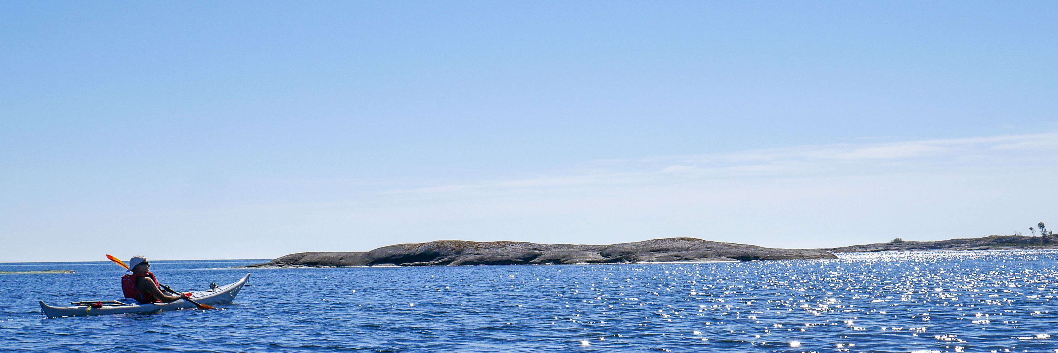 En person paddlar kajak i öppet hav under klarblå himmel med avlägsna öar i bakgrunden.