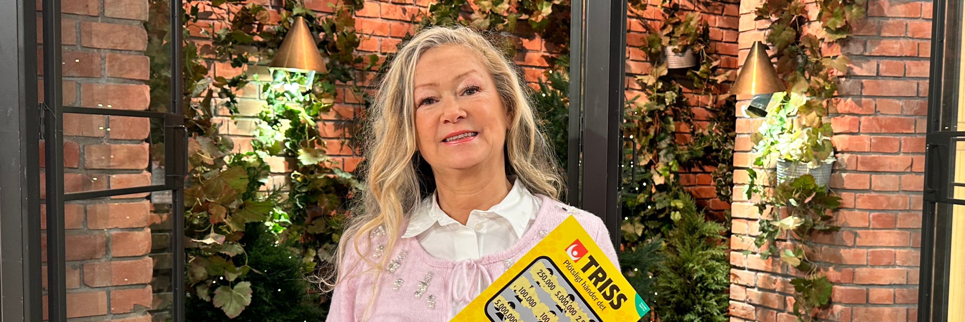 Person med gul Trisslott står framför en bakgrund av tegelväggar och växter.