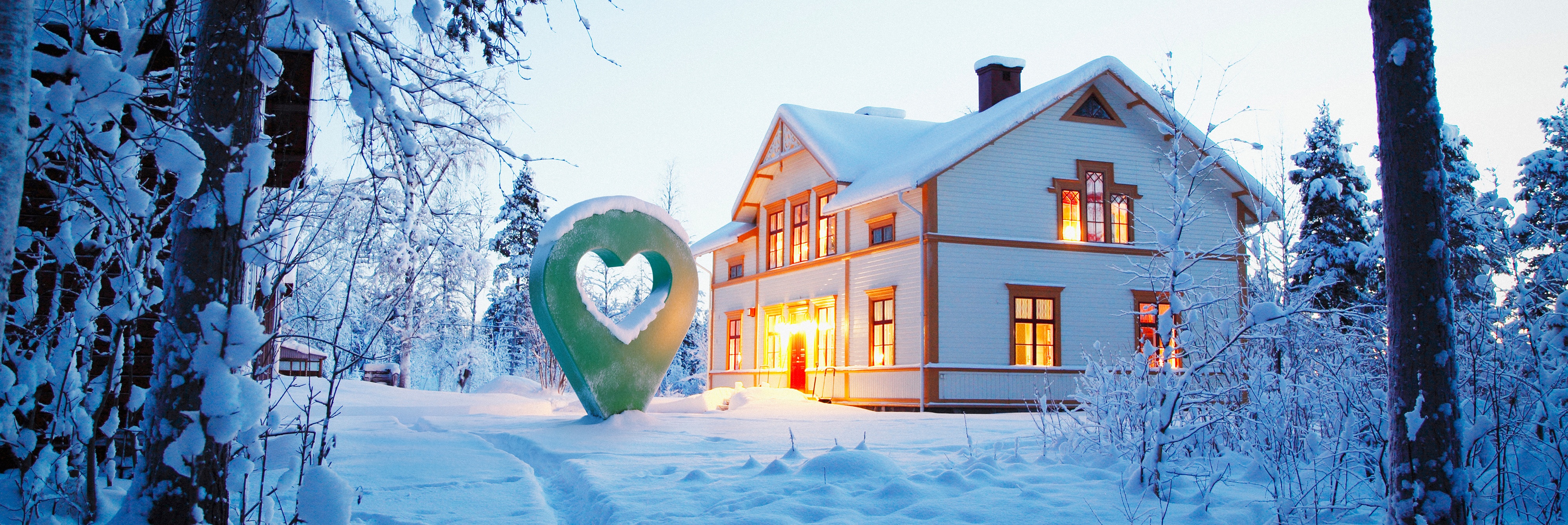 Vackert trähus omgiven av vinterlandskap med snö och en belyst lyckoplatssymbol i skymningen.
