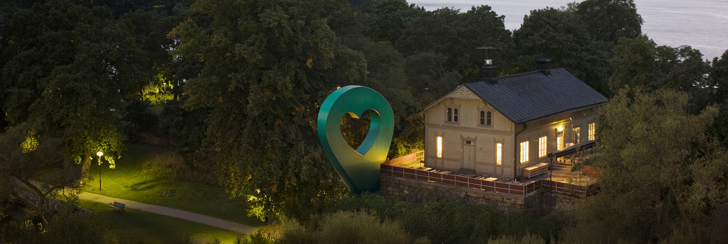 Bild av Lyckplatsen som visar ett hus med ett hjärtaformad skulptur och vacker utsikt mot vattnet i skymningen.