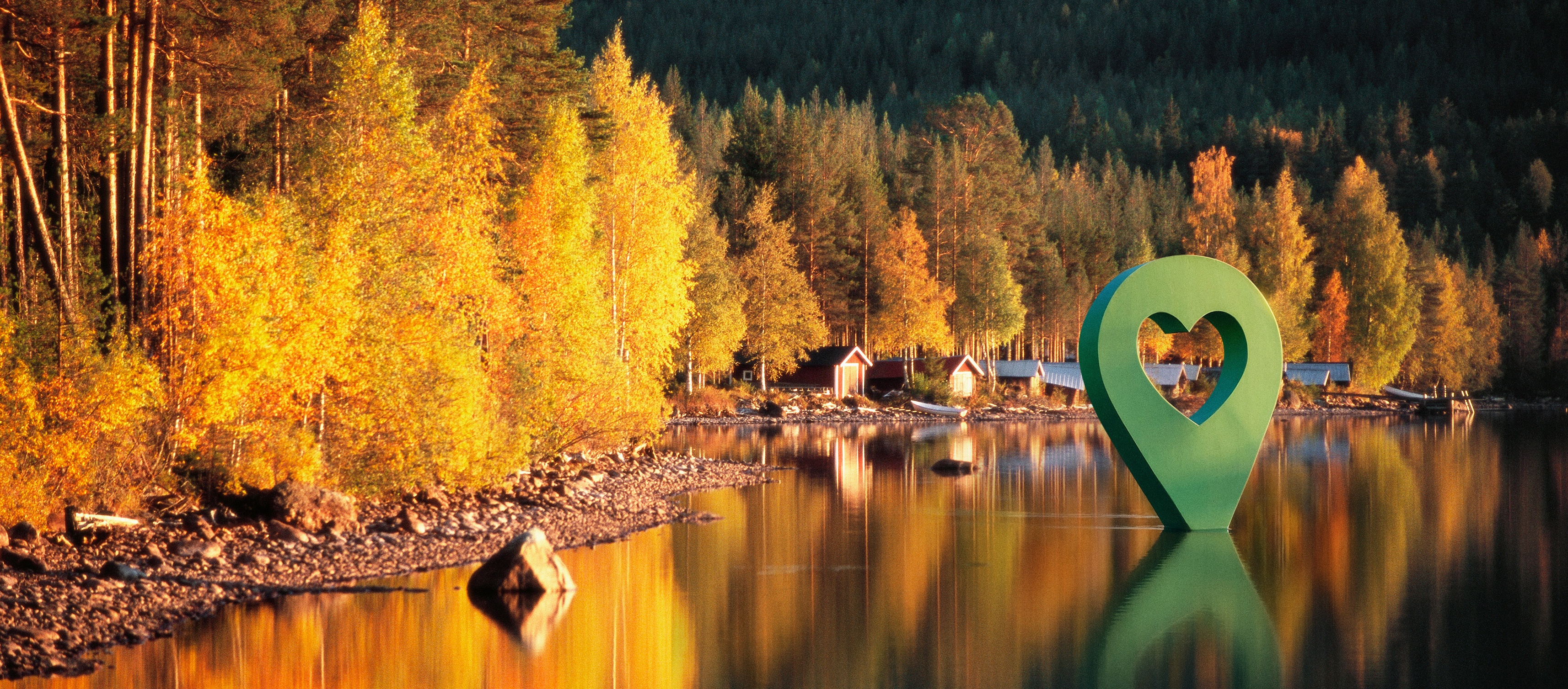 Bild av Lyckplatsen med höstfärger, omringad av träd och en grön hjärtformad markör vid sjön.