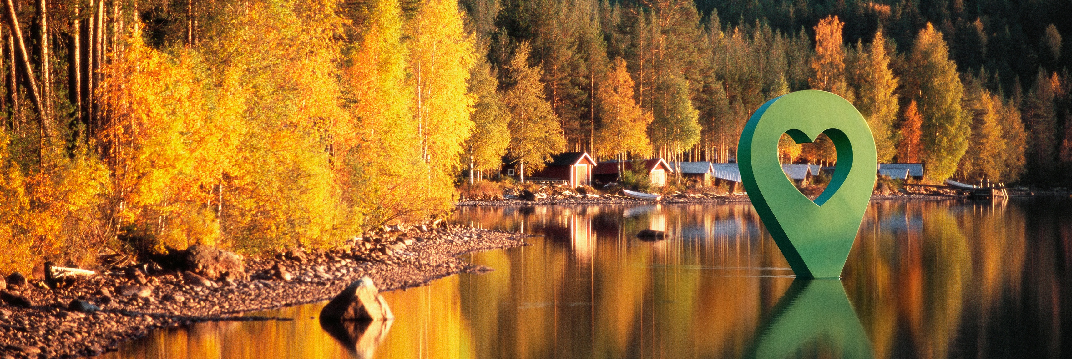 Bild av Lyckplatsen med höstfärger, omringad av träd och en grön hjärtformad markör vid sjön.