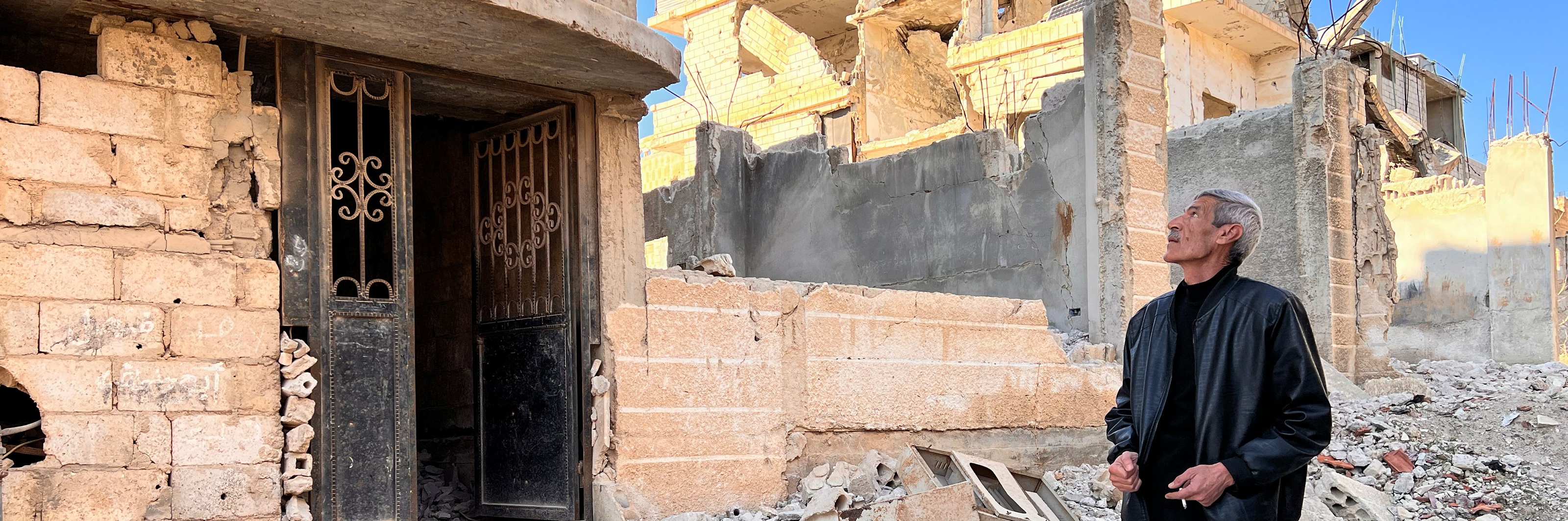 En person står bland ruiner i ett förstört stadsområde i Syrien, med trasiga byggnader omkring.