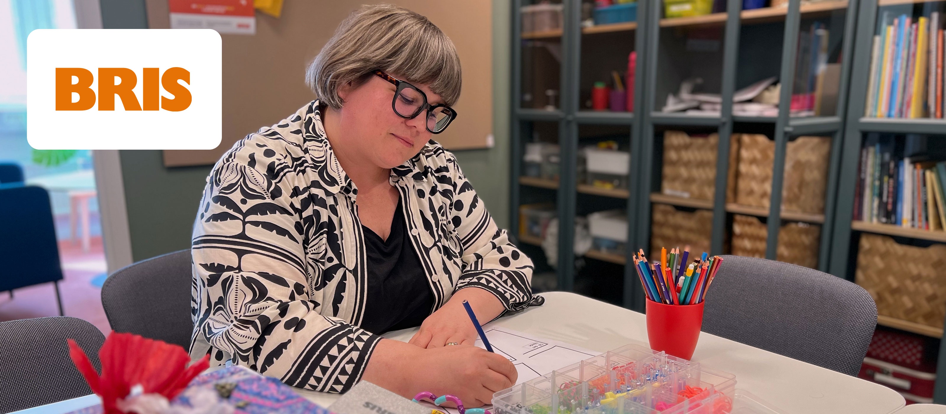 Person sitter vid ett bord och skriver, omgiven av färgglada pennor och material. BRIS-logga synlig.