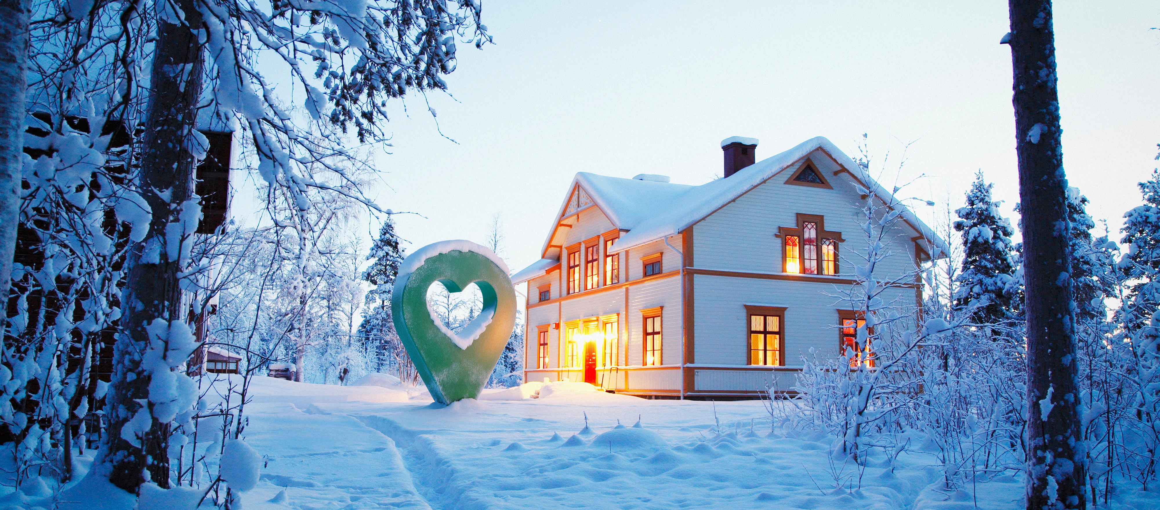 Vackert trähus omgiven av vinterlandskap med snö och en belyst lyckoplatssymbol i skymningen.